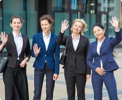 Groep professionele zakenvrouwen die buiten zwaaien voor een modern kantoorgebouw, wat teamwork, zelfvertrouwen en zakelijke samenwerking vertegenwoordigt.